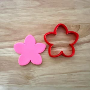 May include: A pink flower-shaped cookie cutter next to a pink flower-shaped cookie. The cookie cutter is red and has a five-petal design. The cookie is a solid pink color. Both are on a wooden surface.