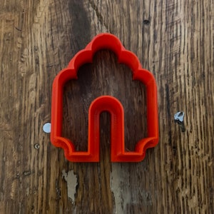 May include: Orange plastic cookie cutter shaped like a house with a rounded roof and a single archway.