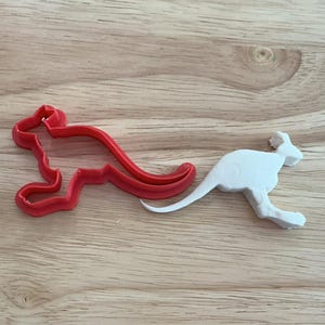 May include: A red plastic kangaroo-shaped cookie cutter next to a white kangaroo-shaped object. The cookie cutter is a silhouette of a kangaroo. The white object is a kangaroo figurine. Both are on a light brown wooden surface.