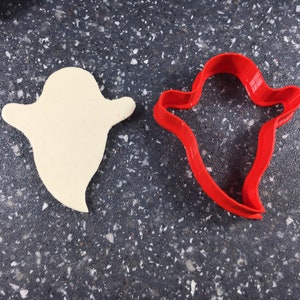 May include: A white ghost cookie cutter and a red ghost cookie cutter on a gray speckled surface.