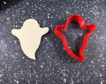 May include: A white ghost cookie cutter and a red ghost cookie cutter on a gray speckled surface.