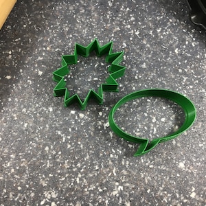 May include: Two green plastic cookie cutters, one shaped like a starburst and the other shaped like a speech bubble.