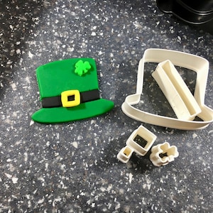 Può includere: Cappello da folletto in pasta di zucchero verde con una fibbia gialla e un trifoglio verde in cima, accanto a uno stampino per biscotti in plastica bianca a forma di cappello da folletto, e due stampini più piccoli, uno a forma di trifoglio e uno a forma di quadrato.