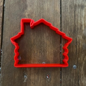 May include: A red plastic cookie cutter shaped like a log cabin. The cutter has a detailed design with a chimney and log details.