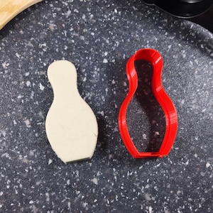 Puede incluir: Un cortador de galletas de plástico rojo en forma de bolos y una galleta blanca con forma de bolos sobre una superficie gris oscura moteada.