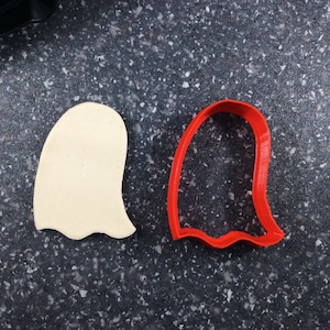 Puede incluir: Una galleta beige con forma de fantasma junto a un cortador de galletas rojo con forma de fantasma. El cortador de galletas es un simple contorno de un fantasma. La galleta está hecha de una masa de color claro. Los artículos están sobre una superficie gris oscuro moteada.