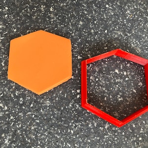 Op de afbeelding: Twee zeshoekige koekjesvormen, een oranje en een rode. De oranje vorm is massief, terwijl de rode vorm hol is.