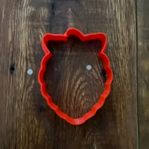 May include: A red plastic cookie cutter shaped like a strawberry. The cutter has a scalloped edge and a leaf on top.