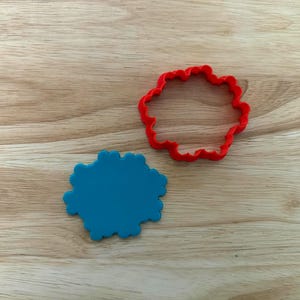 Peut inclure: Un emporte-pièce en plastique rouge et un biscuit bleu. L'emporte-pièce a la forme d'une fleur aux bords arrondis. Le biscuit bleu a également la forme d'une fleur.