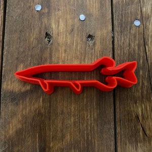May include: A red plastic cookie cutter shaped like a fish. The fish has a detailed design with fins and scales.