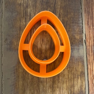 May include: Orange plastic cookie cutter shaped like an egg with a smaller egg-shaped cutout inside.
