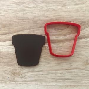 Puede incluir: Un cortador de galletas en forma de maceta marrón oscuro y un cortador de galletas en forma de maceta roja se muestran sobre una superficie de madera. El cortador marrón oscuro es sólido, mientras que el rojo es un contorno.