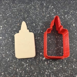 May include: A red plastic cookie cutter shaped like a bottle of glue. The cutter is on a gray speckled surface.