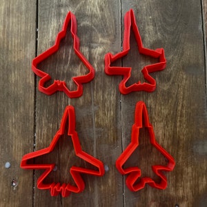 Puede incluir: Cuatro cortadores de galletas de plástico rojo con forma de aviones de combate. Los cortadores están sobre una superficie de madera.