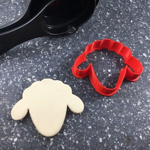 May include: A red plastic cookie cutter shaped like a sheep's head, next to a white cookie cut out in the same shape.