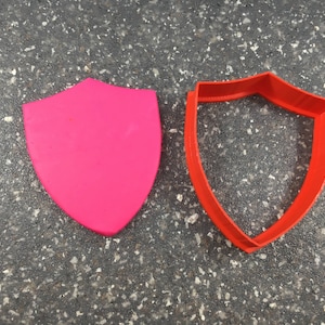 Könnte beinhalten: Ein rosafarbener Schild-Ausstecher aus Plastik und ein roter Schild-Ausstecher aus Plastik.