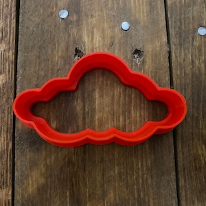 May include: A red plastic cookie cutter shaped like a cloud. The cutter is on a wooden surface.
