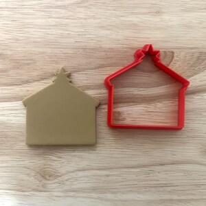May include: Two house-shaped cookie cutters. One is a solid tan color, and the other is a red outline. Both have a star on top. The cutters are on a light brown wooden surface.
