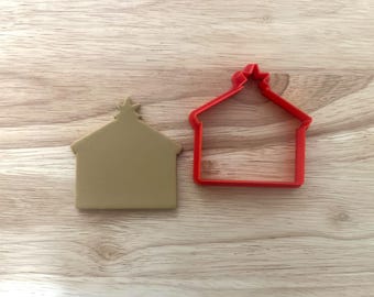 May include: Two house-shaped cookie cutters. One is a solid tan colour, and the other is a red outline. Both have a star on top. The cutters are on a light brown wooden surface.