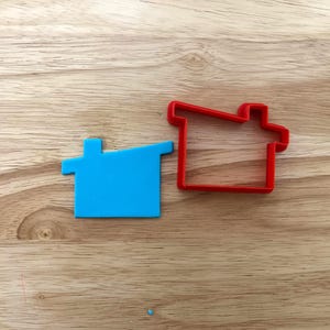May include: Two house-shaped cookie cutters on a wooden surface. One is a solid, bright blue plastic, and the other is a red outline. The cutters are designed for baking and crafting, perfect for creating themed treats or decorations.