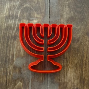 Könnte beinhalten: Ein orangefarbener Ausstecher in Form einer Menora, eines neunarmigen Leuchters. Der Ausstecher ist vor einem rustikalen Holzhintergrund platziert, der sein Design zum Backen hervorhebt. Die Menora ist ein Symbol des jüdischen Glaubens.