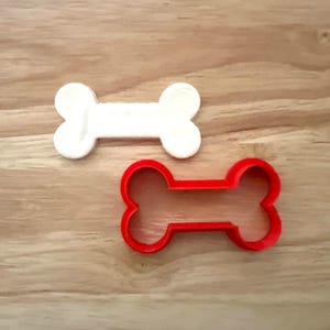 以下が含まれることがあります： 2つの犬の骨の形をしたオブジェクト。1つは白で、もう1つは赤で輪郭です。どちらも明るい茶色の木製の表面にあります。これらはクッキーカッターまたはクラフト用品である可能性があります。