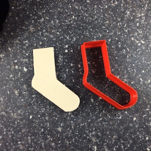 May include: A red plastic cookie cutter shaped like a sock, next to a white sock-shaped cookie cutter.
