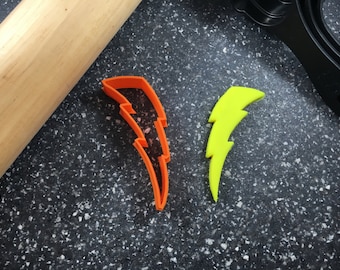 Puede incluir: Un cortador de galletas naranja con forma de rayo y un rayo amarillo se muestran sobre una superficie moteada. También se ven un rodillo de madera y un utensilio de cocina negro.
