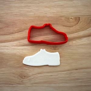 May include: A red plastic cookie cutter in the shape of a sneaker sits above a white sugar cookie in the same shape. The objects are on a wooden surface.