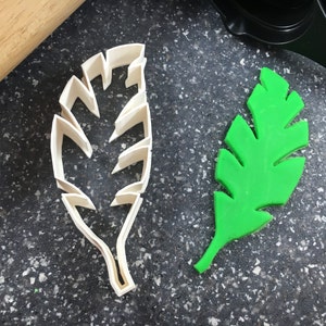 Puede incluir: Un cortador de galletas de plástico blanco en forma de hoja y una hoja de fondant verde. El cortador de galletas está detallado con múltiples líneas y la hoja de fondant es una forma sólida.