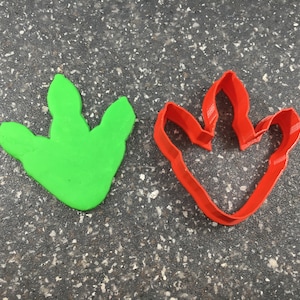 May include: A green dinosaur footprint made of play dough next to a red plastic dinosaur footprint cookie cutter.