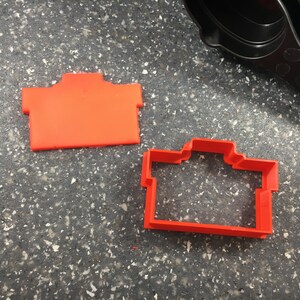 May include: Two red plastic cookie cutters shaped like a house. The cutter on the left is solid, while the cutter on the right is hollow.