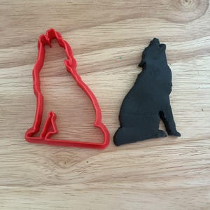 Puede incluir: Un cortador de galletas de plástico rojo con forma de lobo junto a una galleta negra con forma de lobo. El lobo está aullando. Los artículos están sobre una superficie de madera de color marrón claro. El cortador es un solo contorno.