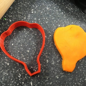 May include: A red plastic cookie cutter shaped like a hot air balloon, next to a yellow piece of modeling clay cut out with the cookie cutter.