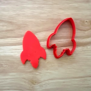 May include: Two red rocket-shaped cookie cutters on a light wood grain surface. One cutter is a solid rocket shape, and the other is the outline of a rocket. The cutters are likely for baking or crafting.