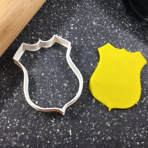 May include: A white plastic cookie cutter shaped like a police badge, next to a yellow cookie dough cutout of the same shape.