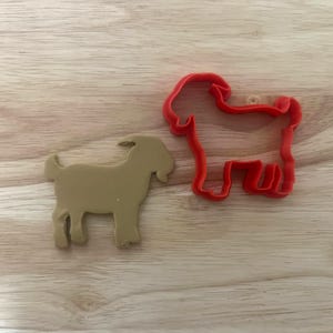 Könnte beinhalten: Ein hellbrauner Ziegen-Ausstecher und ein roter Ziegen-Ausstecher liegen auf einer hellen Holzoberfläche. Der hellbraune Keks hat die Form einer Ziege. Der rote Ausstecher ist eine Ziegenkontur.