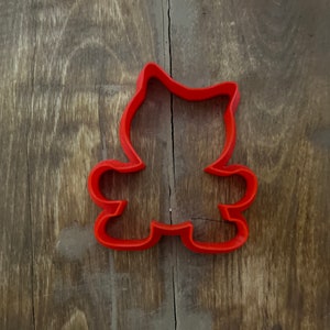 May include: Red plastic cookie cutter shaped like a cartoon bear sitting down with its arms outstretched.