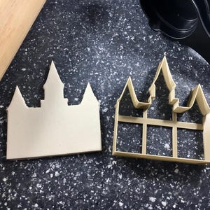 Pode incluir: Um cortador de biscoitos de plástico branco em forma de castelo com um design detalhado. O cortador está ao lado de um biscoito em branco que foi cortado com o cortador.