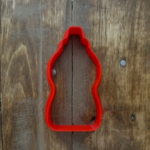 May include: Red plastic cookie cutter shaped like a bottle. The cutter is on a wooden surface.