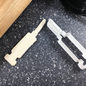 May include: Two white plastic cookie cutters shaped like syringes. The cutters are on a black speckled surface.