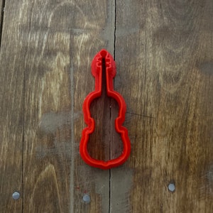 Pode incluir: Um cortador de biscoitos em forma de violino vermelho brilhante. O cortador é feito de plástico e tem um contorno detalhado de um violino. O cortador está sobre um fundo de madeira rústica.