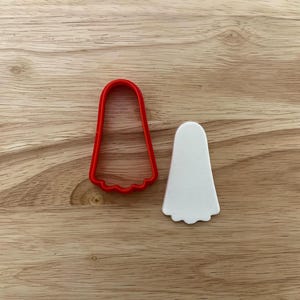 May include: A red plastic ghost-shaped cookie cutter next to a white ghost-shaped cookie. The cutter has a scalloped bottom edge. The cookie is also ghost-shaped with a scalloped bottom edge. The items are on a wooden surface.