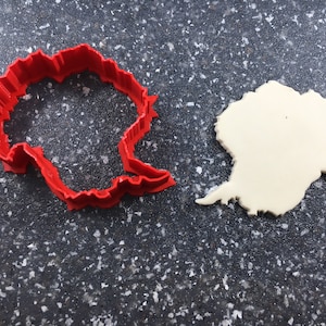 May include: A red plastic cookie cutter shaped like the continent of Antarctica, next to a white cookie dough cutout of the same shape.