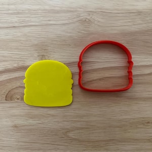 May include: A yellow plastic burger patty and a red plastic burger bun cookie cutter sit on a wooden surface. The patty is a solid yellow color, and the bun cutter is a solid red color. The items are for baking.
