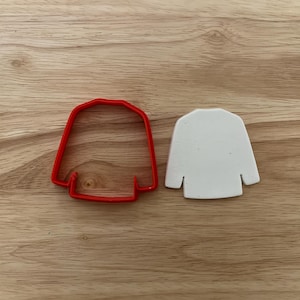 May include: A red plastic sweater-shaped cookie cutter next to a white sweater-shaped cookie. The cookie cutter is made of red plastic and has a simple outline. The white cookie is smooth and has a similar shape.