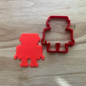 May include: Two red robot-shaped cookie cutters sit on a wooden surface. One is a solid shape, and the other is the outline of the same robot. The cutters have a simple, blocky design, perfect for making robot-themed cookies or treats.