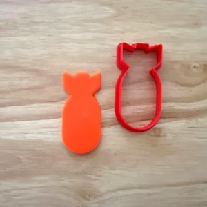 May include: Two bomb-shaped cookie cutters. One is solid orange, the other is a red outline. Both have a rounded body and a notched top. They are placed on a light brown wooden surface.