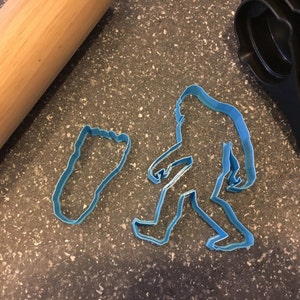May include: Blue plastic cookie cutters shaped like a Bigfoot and a footprint. The cutters are on a gray speckled surface.