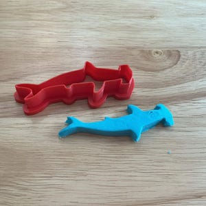 Könnte beinhalten: Ein roter Kunststoff-Hammerhai-Ausstecher neben einem blau geformten Hammerhai-Keks. Der Ausstecher ist eine solide Umrisslinie des Hais. Der Keks hat eine leuchtend blaue Farbe.
